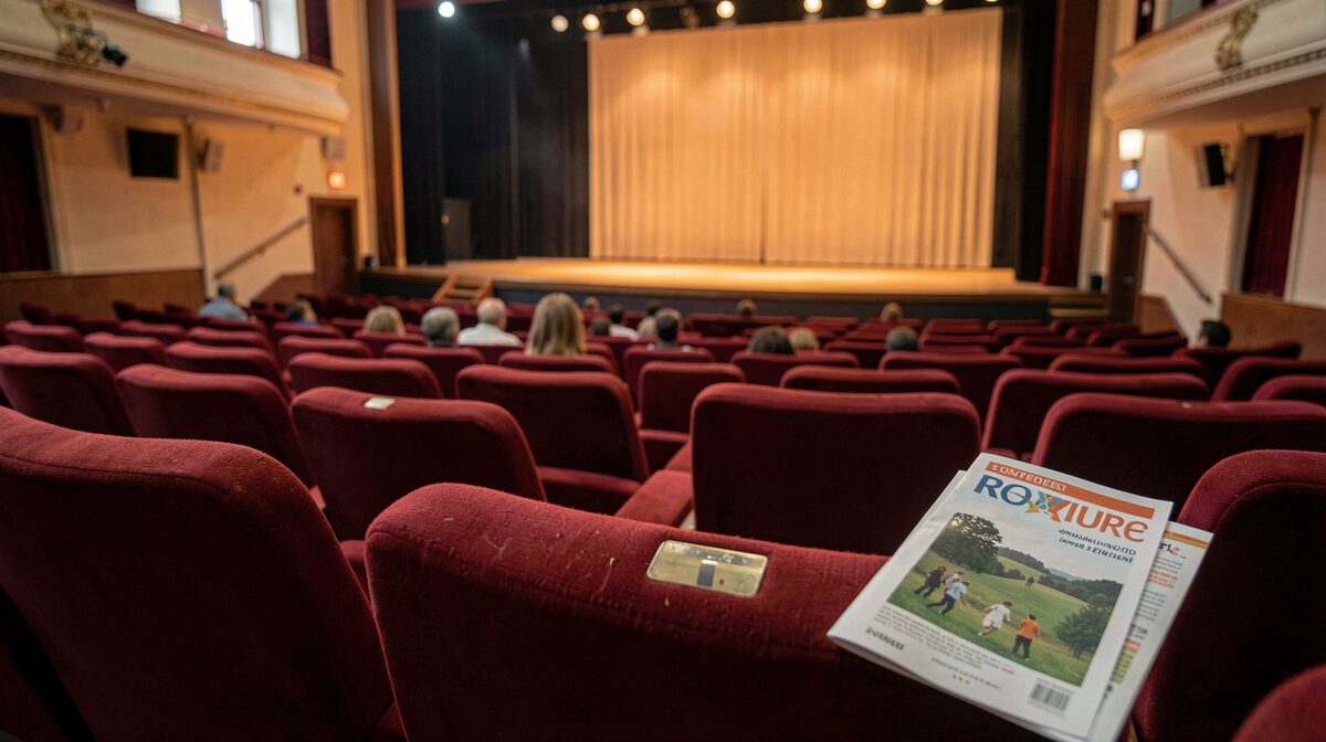 Een lege theaterzaal met rode stoelen, warm licht op het podium, programmafolder op een stoel in de voorgrond, vanuit de zaal gefotografeerd.